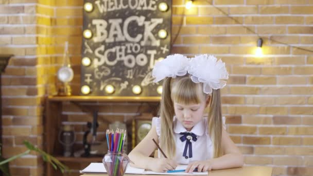 Cute little girl siedzi przy stole i rysuje z kolorowych ołówków na tle klasy szkolnej. W lekcji uczennica z białymi kokardkami pisze ołówkami.. — Wideo stockowe