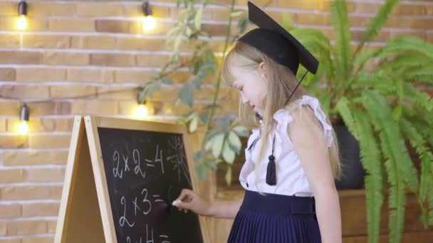 Meisje schrijft met krijt op schoolbord op achtergrond van de schoolklas. Een basisschoolmeisje met een zwarte muts. Een schoolmeisje leert vermenigvuldiging in de eerste rekenles. — Stockvideo