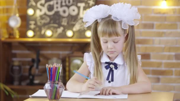 Linda niña se sienta a la mesa y dibuja con lápices de colores en el fondo de una clase de la escuela. Una niña de primaria con lazos blancos escribe con lápices en la lección. De cerca.. — Vídeo de stock