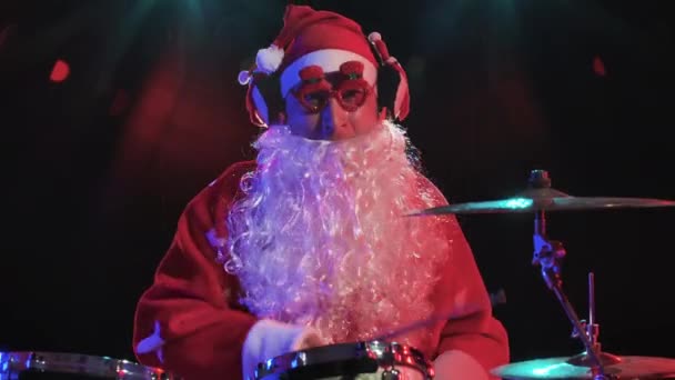 Le Père Noël en costume rouge avec barbe et lunettes amusantes joue de la batterie dans un studio sombre avec des lumières vives. Un homme en costume de carnaval exécute un spectacle lors d'une fête du Nouvel An. Ferme là. Mouvement lent. — Video