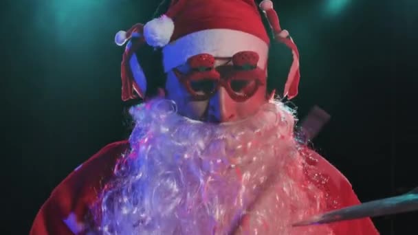 Le Père Noël en costume rouge avec barbe et lunettes amusantes joue de la batterie dans un studio sombre avec des lumières vives. Un homme en costume de carnaval exécute un spectacle lors d'une fête du Nouvel An. Ferme là. Mouvement lent. — Video