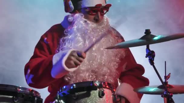 Babbo Natale in costume rosso con barba e bicchieri divertenti suona la batteria in studio scuro con luci brillanti e fumo. Un uomo in costume da carnevale esegue uno spettacolo a una festa di Capodanno. Chiudete. Rallentatore. — Video Stock