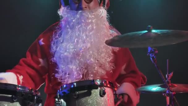 Babbo Natale in costume rosso con barba e bicchieri divertenti suona la batteria in uno studio buio con luci brillanti. Un uomo in costume da carnevale fa uno spettacolo a una festa di Capodanno. Chiudete. Rallentatore. — Video Stock