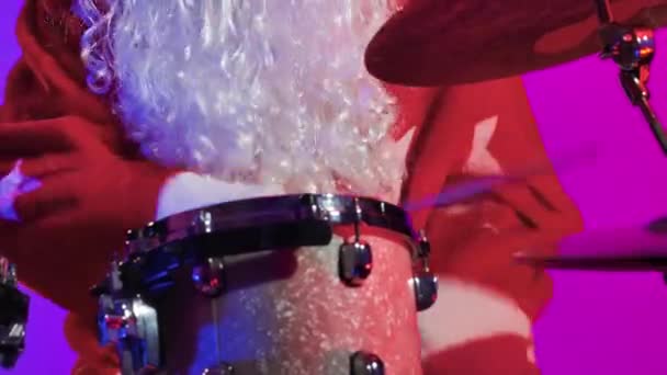 En man klädd som jultomten spelar en trumset i en mörk studio mot en bakgrund av ljusa lila ljus. En musiker i karnevalsdräkt uppträder på julfesten. Närbild. Långsamma rörelser. — Stockvideo