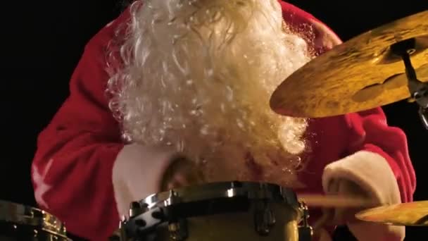 Músico alegre vestido como Santa Claus toca la batería en un fondo de estudio negro. Un hombre con un traje rojo festivo con barba golpea tambores y platillos de percusión con palos. De cerca. Movimiento lento. — Vídeos de Stock