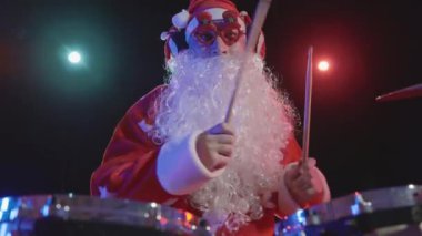 Noel Baba 'nın şenlikli performansı. Yeni yıl kostümü giymiş sakallı bir adam davul setini canlı çalıyor, aktif olarak davul ve perküsyon zillerini sopasıyla vuruyor. Kapatın. Yavaş çekim.