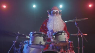Kırmızı sakallı, komik gözlüklü Noel Baba, parlak ışıklarla dolu karanlık bir stüdyoda davul çalıyor. Karnaval kostümü giymiş bir adam yeni yıl partisinde gösteri yapıyor. Yavaş çekim.