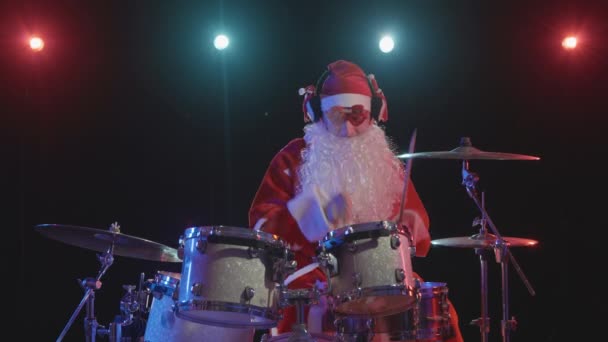 Санта-Клаус в красном костюме с бородой и смешными очками играет на барабанах в темной студии с яркими огнями. Человек в карнавальном костюме выступает с шоу на новогодней вечеринке. Медленное движение. — стоковое видео