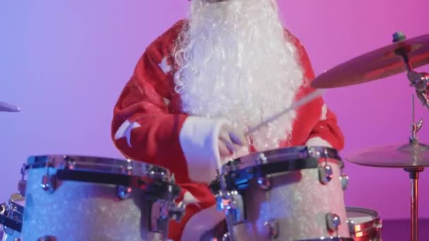 サンタクロースの格好をした男が、明るい紫色の光を背景に暗いスタジオでドラムキットを演奏する。カーニバルの衣装を着た音楽家がクリスマスパーティーで演奏する。閉めろ。スローモーション. — ストック動画