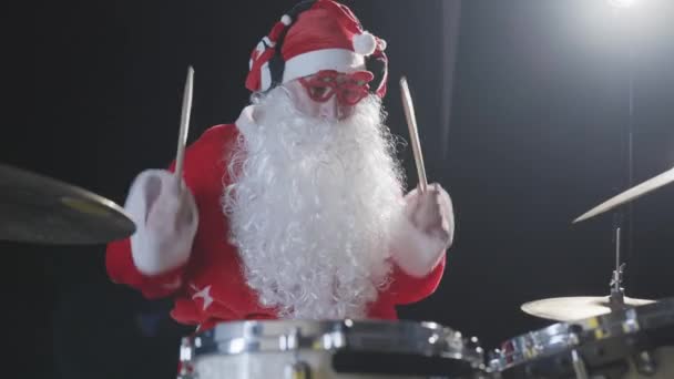 Musicista allegro vestito come Babbo Natale suona la batteria su uno sfondo retroilluminato studio nero. Un uomo in abito rosso festivo con la barba colpisce tamburi e piatti a percussione con bastoni. Rallentatore. — Video Stock