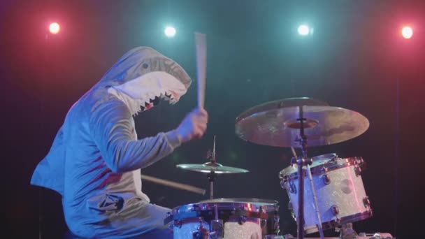 Vue latérale du musicien en costume de requin festif joue de la batterie et frappe des cymbales à percussion. Un musicien joue lors d'un concert festif dans le noir avec des lumières vives et de la fumée. Mouvement lent. — Video