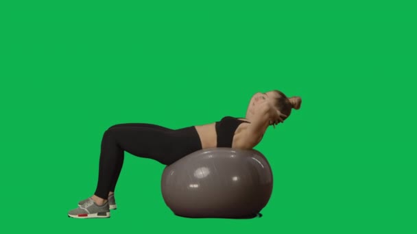 Eine junge Frau in Sportbekleidung schüttelt die Presse am Fitnessball. Bauchmuskeltraining, Fitness für Frauen. Seitenansicht in voller Länge auf grünem Hintergrund. Zeitlupe bereit 59.94fps. — Stockvideo