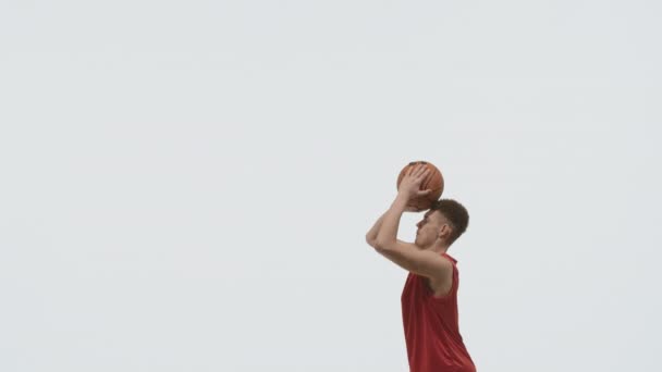 バスケットボールの男はバスケットにボールを投げ、彼が得点したことを嬉しく思います。白いスタジオを背景にしたスポーツユニフォームを着た若いスポーツマン。スローモーション. — ストック動画