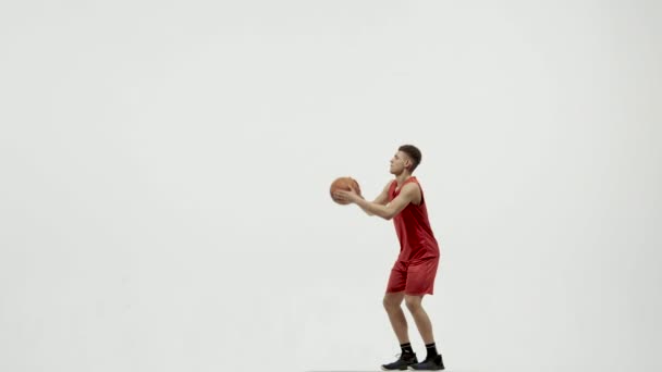 El jugador de baloncesto lanza la pelota a la canasta y se alegra de haber anotado. Joven deportista con uniforme deportivo entrena sobre un fondo blanco de estudio. Movimiento lento. — Vídeo de stock