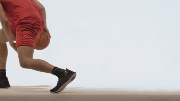 Geschicktes Dribbling eines Basketballspielers im Studio auf weißem Hintergrund. Ein großer junger Sportler in roter Sportuniform schlägt den Ball auf den Boden. Aus nächster Nähe. Zeitlupe bereit 59.94fps. — Stockvideo