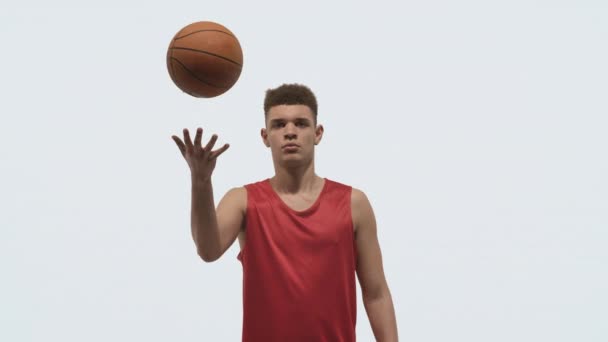 Porträt eines jungen Basketballspielers, der in die Kamera schaut und den Ball nach oben wirft. Ein Athlet in roter Sportuniform posiert vor weißem Studiohintergrund. Aus nächster Nähe. Zeitlupe bereit 59.94fps. — Stockvideo
