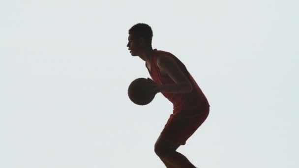 Vista lateral ou jogador de basquete jovem em corridas uniformes de esportes vermelhos e dribles em fundo estúdio branco. O atleta está se preparando para a competição em treinamento. Silhueta. Movimento lento pronto 59.94fps. — Vídeo de Stock