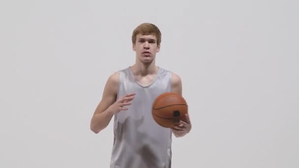 Genç basketbolcunun portresi kameraya bakıyor ve elden ele top atıyor. Spor kıyafetleri içinde beyaz stüdyo geçmişine karşı poz veren bir sporcu. Kapatın. Yavaş çekim hazır 59.94fps. — Stok video