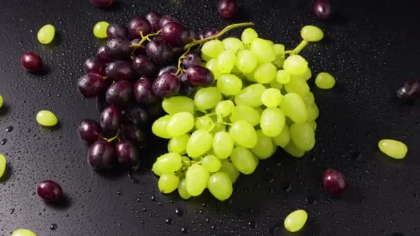 Na mokrej powierzchni stołu leżą pęczki zielonych i niebieskich winogron z kroplami wilgoci. Widok z góry zbliżenie dojrzałych jagód winogron obracających się w zwolnionym tempie. Winorośl w kroplach wilgoci. Słodkie owoce. — Wideo stockowe