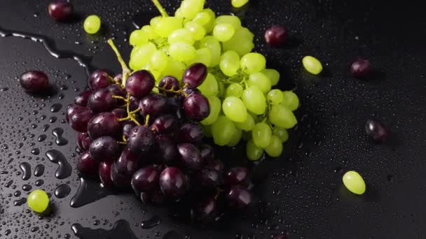 Des grappes de raisins verts et bleus avec des gouttes d'humidité se trouvent sur la surface de la table humide. Vue de dessus gros plan de baies de raisin mûres tournant au ralenti. La vigne dans des gouttes d'humidité. Fruits doux. — Video