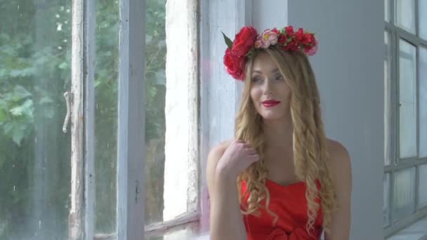 Mujer joven y bonita con corona de flores rojas en la cabeza sentada en el alféizar de la ventana, sonriendo, en cámara lenta — Vídeos de Stock