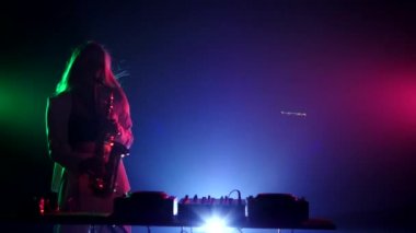Young sexy, blonde woman dj, bending, in white jacket and black top playing music using saxophone, flipping hair, dancing,  on green, pink and blue, backlight