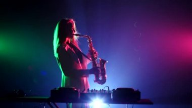Young sexy, blonde woman dj in white jacket and black top playing music using saxophone, flipping hair, dancing, on green, pink and blue, backlight
