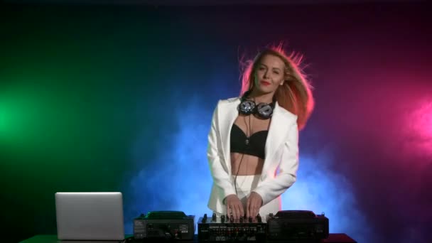 Hermosa, encantadora y sonriente chica dj en chaqueta blanca, auriculares con música y baile, humo — Vídeos de Stock