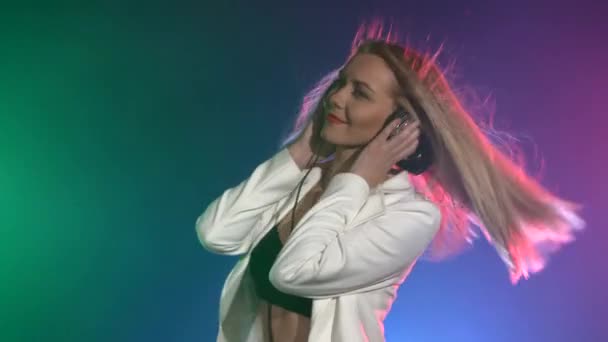 Hermosa, encantadora y sonriente chica dj con chaqueta blanca, auriculares en sus oídos, tocando, escuchando música y bailando, fumando — Vídeos de Stock