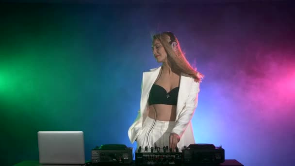 Attraktiv DJ flicka i sexiga kläder i vit jacka och svart topp, med hörlurar, rök, bakgrundsbelysning — Stockvideo