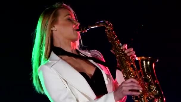 Young sexy, blonde woman dj in white jacket playing music using saxophone on black, cam moves upward — ストック動画