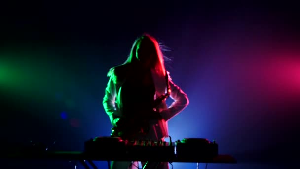 Young sexy, blonde woman dj, in white jacket and black top playing music using saxophone, flipping hair, dancing, hand up  cam moves upwards, on green, pink and blue, backlight — Wideo stockowe