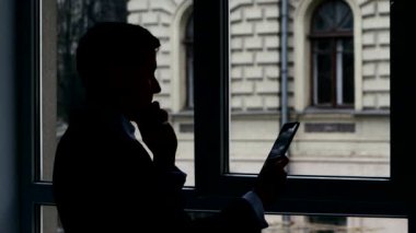 Pencerede duran işadamı, telefondan konuşurken, tabletteki bilgileri görüntüle. Siluet