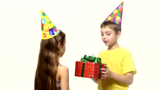 El chico le da un regalo rojo a una chica. Ambos están en gorras festivas sobre fondo blanco — Vídeos de Stock