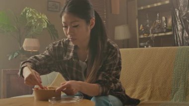 A Korean woman is sitting comfortably on a sofa, using chopsticks to eat Asian noodles from a bowl.