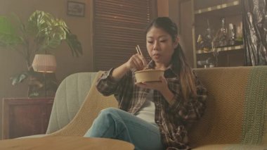 A Korean woman seated on a cozy sofa, savoring Asian food from a bowl using chopsticks in a relaxed setting.