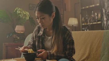 A Korean woman sitting comfortably and eating delicious Asian food with chopsticks in her cozy living room.