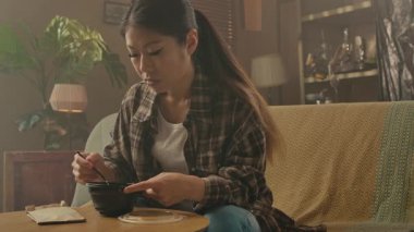 A Korean woman sitting comfortably and eating delicious Asian food with chopsticks in her cozy living room.