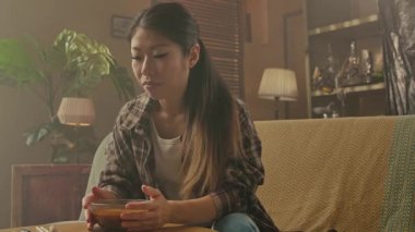 A Korean woman enjoys a bowl of Asian food in a cozy home setting, highlighting cultural dining practices.