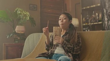 A Korean woman in a casual setting enjoying a piece of Asian food while relaxing on a comfortable sofa at home.
