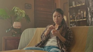 A Korean woman sits on a sofa, casually eating Asian snacks in a cozy and stylish living room.