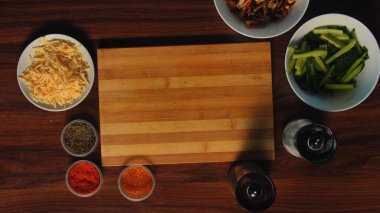 Tahta kesme tahtası. Etrafı peynir, salatalık dilimleri ve ahşap masadaki baharatlarla çevrili..