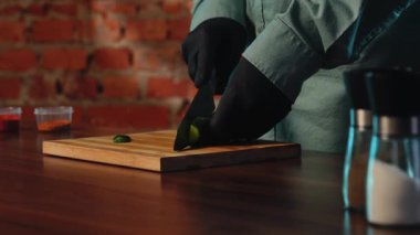 A chef wearing black gloves slices fresh cucumbers on a wooden board in a rustic kitchen setting. The process of preparing food in a cafe or restaurant.