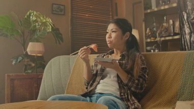 A Korean woman is seated on a couch, savoring Asian food with chopsticks in a cozy home interior.
