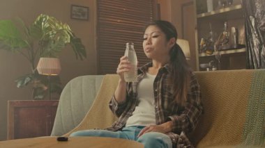 A young woman drinks juice from a plastic bottle. A Korean woman sits on the sofa, enjoying delicious Asian cuisine in a cozy, beautifully decorated room.
