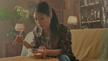 A Korean woman seated on a cozy sofa, savoring Asian food from a bowl using chopsticks in a relaxed setting.