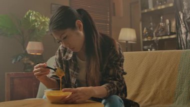 A Korean woman seated on a cozy sofa, savoring Asian food from a bowl using chopsticks in a relaxed setting.