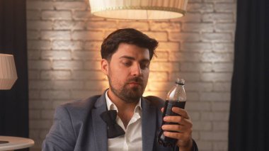 A businessman in a business suit drinks soda from a plastic bottle in a modern room with ambient lighting. An office worker relaxes during lunch.