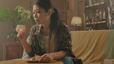 A Korean woman savoring Asian food while sitting on a cozy couch in a warmly lit living room.