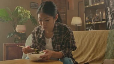A Korean woman is sitting comfortably on a sofa, enjoying Asian noodles with chopsticks in a warmly decorated living room.